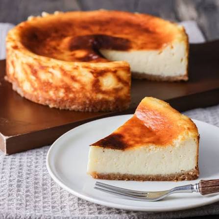 白い皿に盛り付けた一切れのベイクドチーズケーキと、奥に置かれたホールケーキ